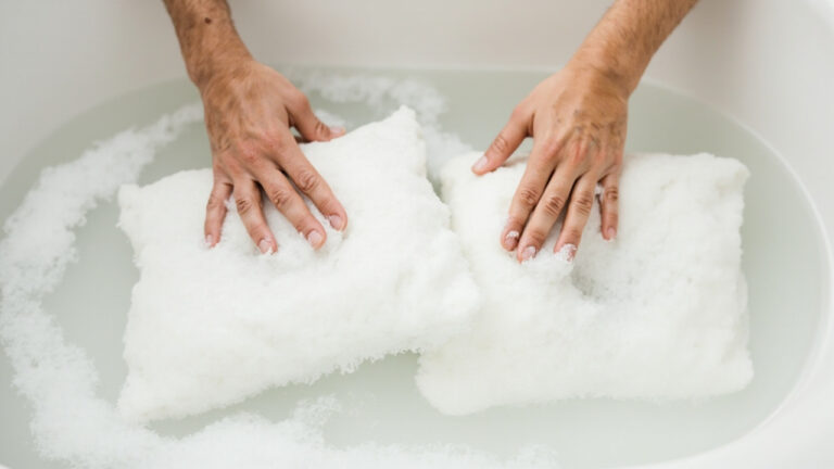 "Hands gently hand-washing a white memory foam pillow in a sink with lukewarm soapy water"